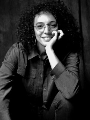 a black and white image of a girl with glasses and curly hair sitting and resting her chin in her hand