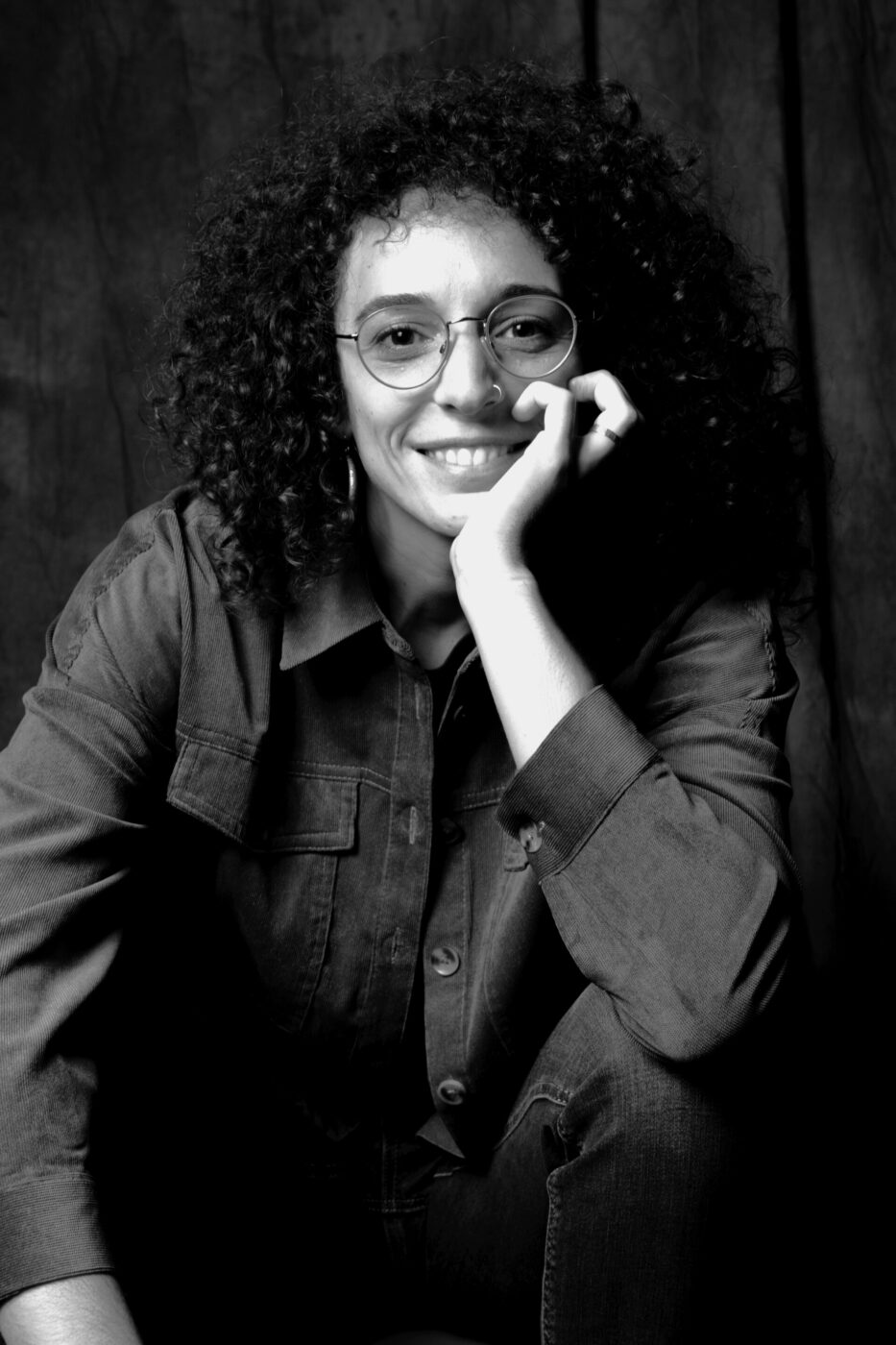 a black and white image of a girl with glasses and curly hair sitting and resting her chin in her hand