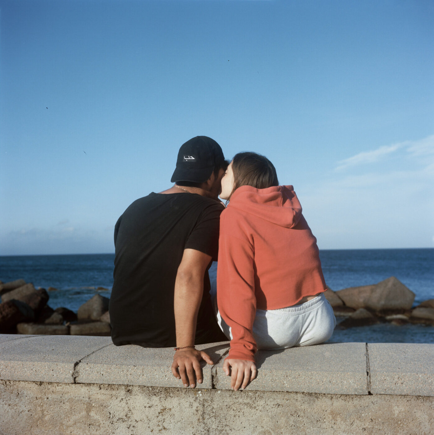 an image of two young teens whispering something to each other photographed from behind sitting along a wall by the sea