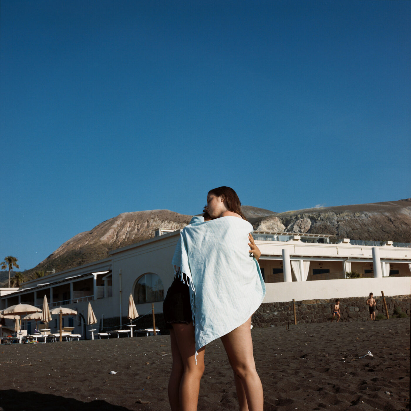 an image of two teens hugging with a beach towel wrapped around them on the gray sand of an aeolian island