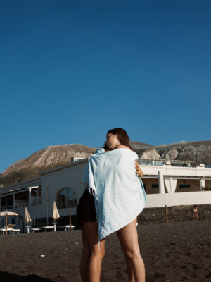 an image of two teens hugging with a beach towel wrapped around them on the gray sand of an aeolian island