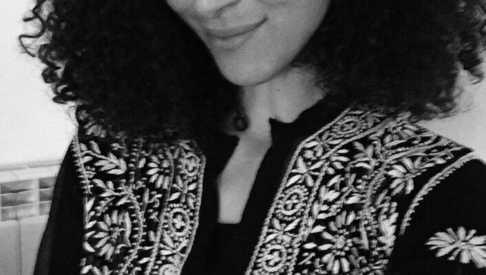 : In questa foto in bianco e nero, una persona con i capelli ricci, che sorride leggermente, indossa un abito tradizionale decorato con ricami floreali dettagliati.