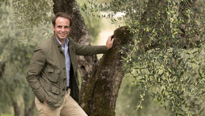 an image of a man in pants and a button up leaning against an olive tree