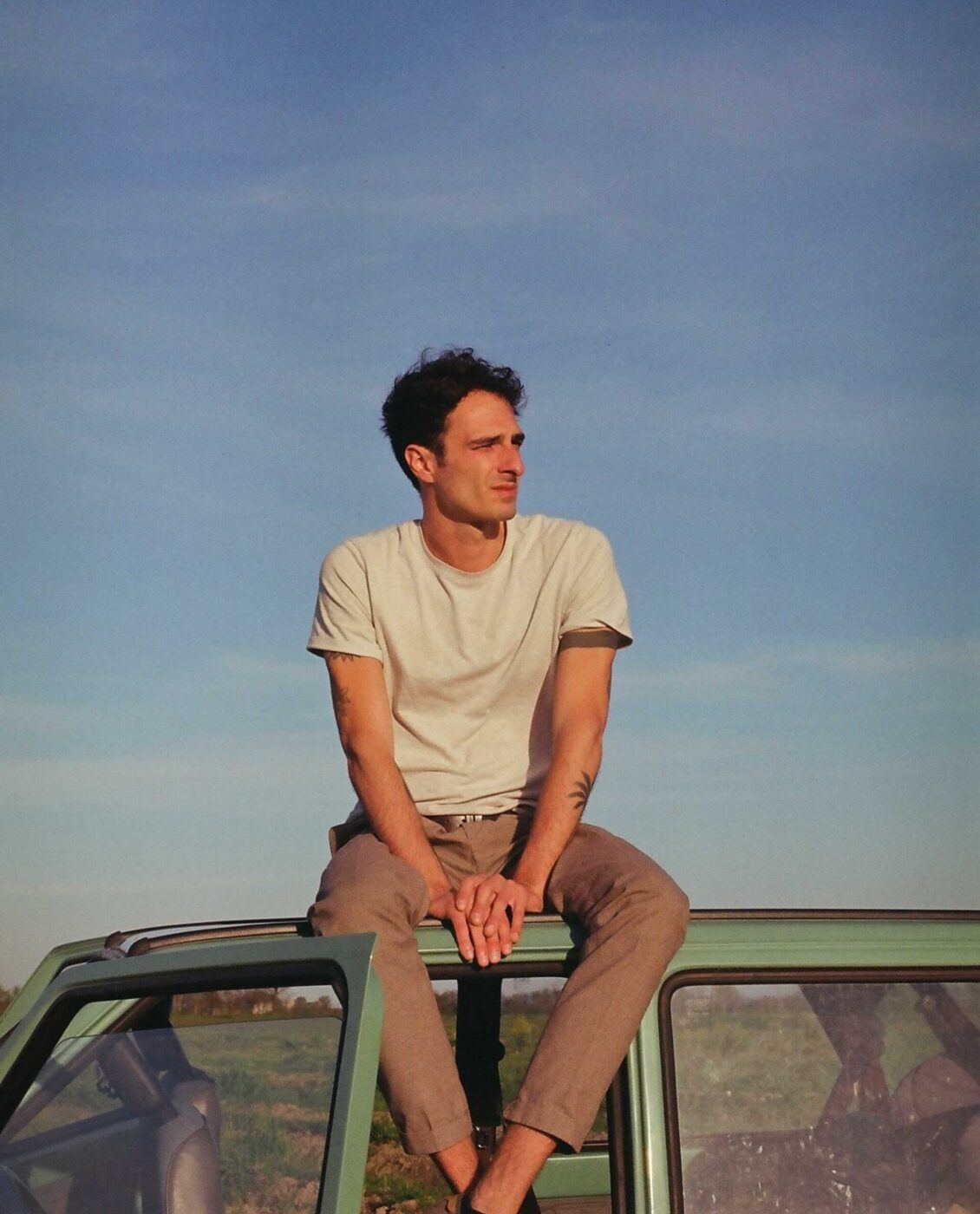 an image of a young man sitting on top of a light green car with a blue sky in the background