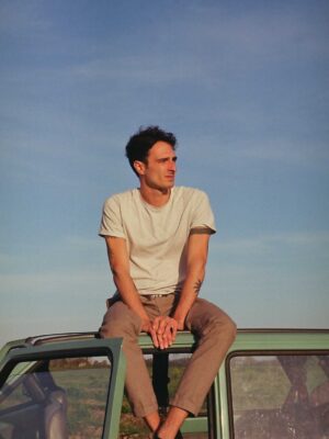 an image of a young man sitting on top of a light green car with a blue sky in the background