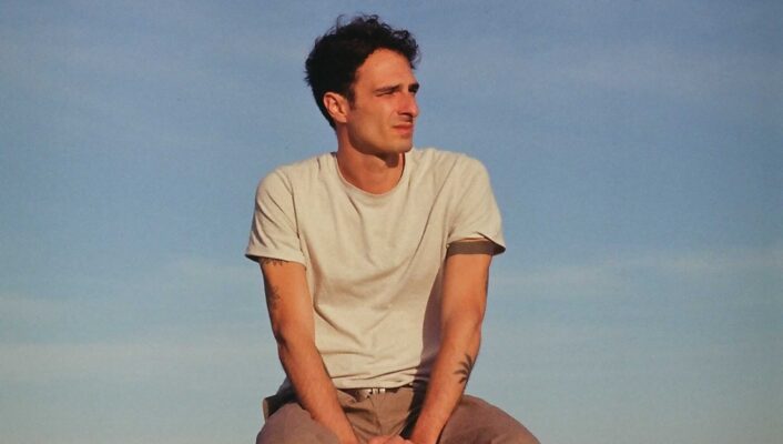 an image of a young man sitting on top of a light green car with a blue sky in the background