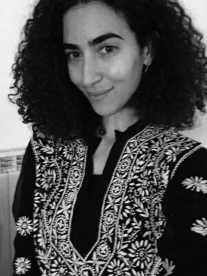Black and white photo of a person with curly hair, wearing a traditional garment with detailed, ornate embroidery. The individual is smiling slightly and facing the camera.