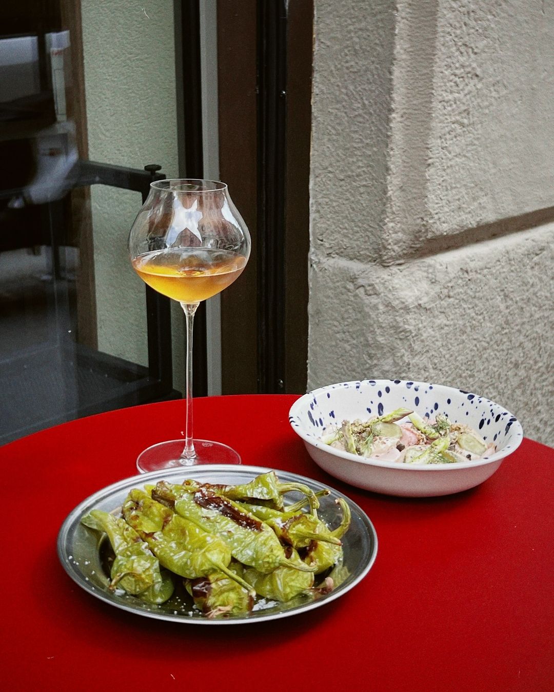 a red table with shishito peppers, a salad, and a glass of orange wine for aperitivo at minerale in milan