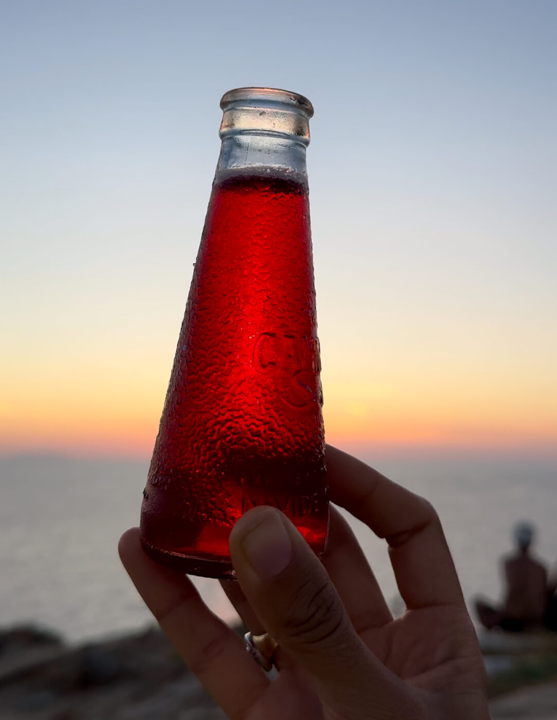 someone holding a bottle of campari soda against a sunset in italy