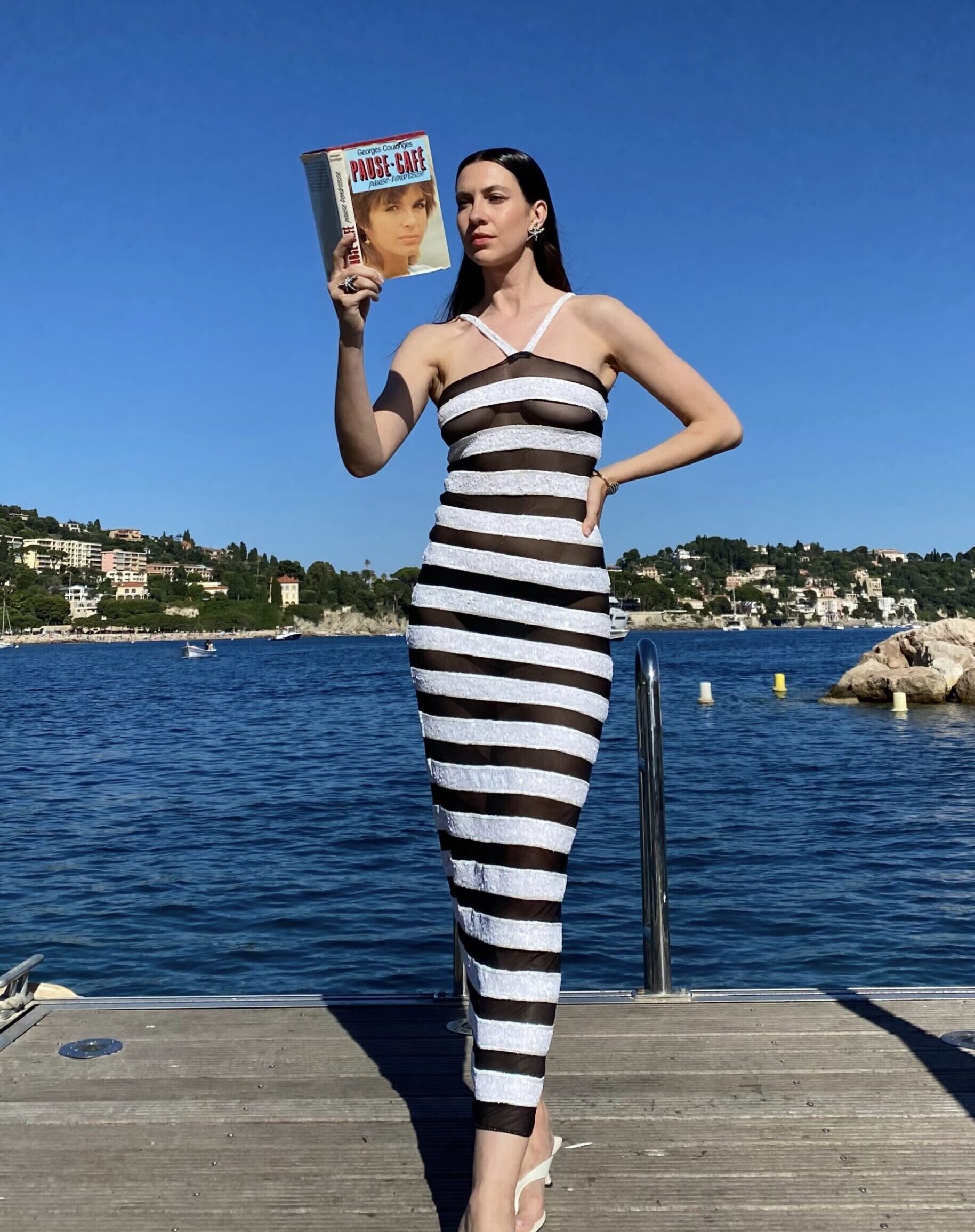 Popa in a stripped see through dress on a dock reading a book