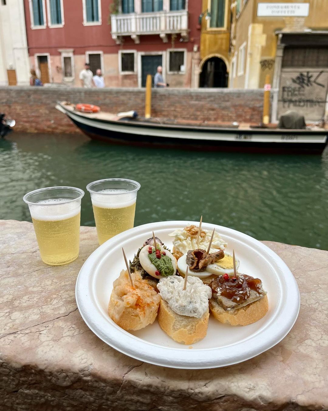 two glasses of sparkling wine in plastic cups and a plastic plate with assorted cicchetti along the canal in venice