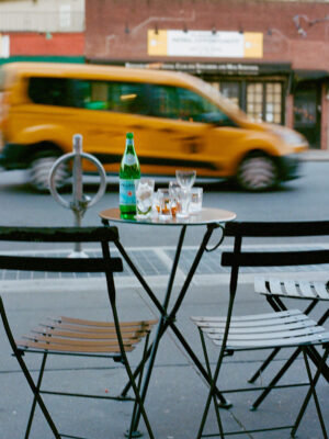 A small café table with two empty chairs, a bottle of sparkling water, and empty glasses sits on a city sidewalk; yellow taxi speeds by.