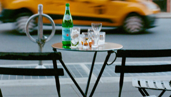 A small café table with two empty chairs, a bottle of sparkling water, and empty glasses sits on a city sidewalk; yellow taxi speeds by.