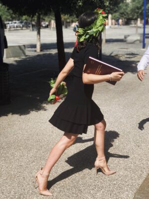 An image of a woman walking away on her graduation day wearing a laurel crown and holding her diploma
