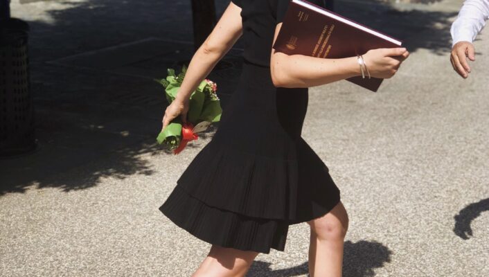 An image of a woman walking away on her graduation day wearing a laurel crown and holding her diploma