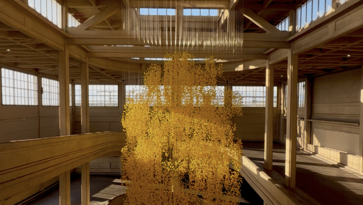 A detailed yellow sculpture hangs from the ceiling of a bright, industrial space with beams and big windows. Sun casts geometric shadows.