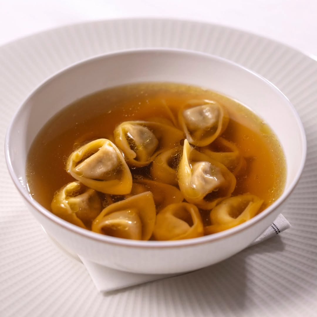 Bowl of tortellini in clear broth, placed on a white plate with a napkin beneath. The tortellini are filled pasta shapes, floating in the golden liquid, set against a simple background.