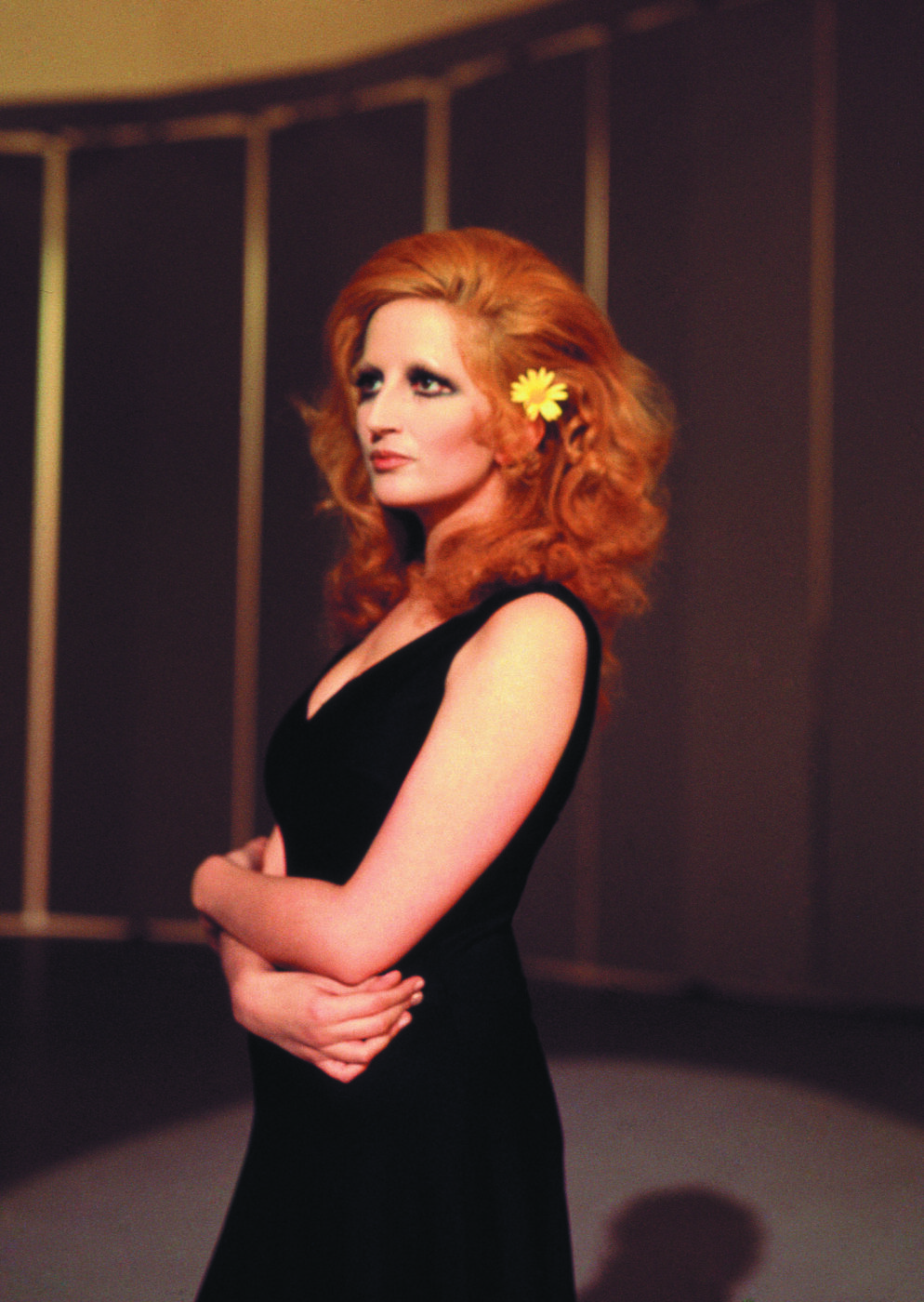 Confident Elegance - Person with long red hair and a yellow flower tucked behind one ear stands confidently, wearing a sleeveless black dress. Arms crossed, they look to the side. The background features vertical lines and warm lighting.