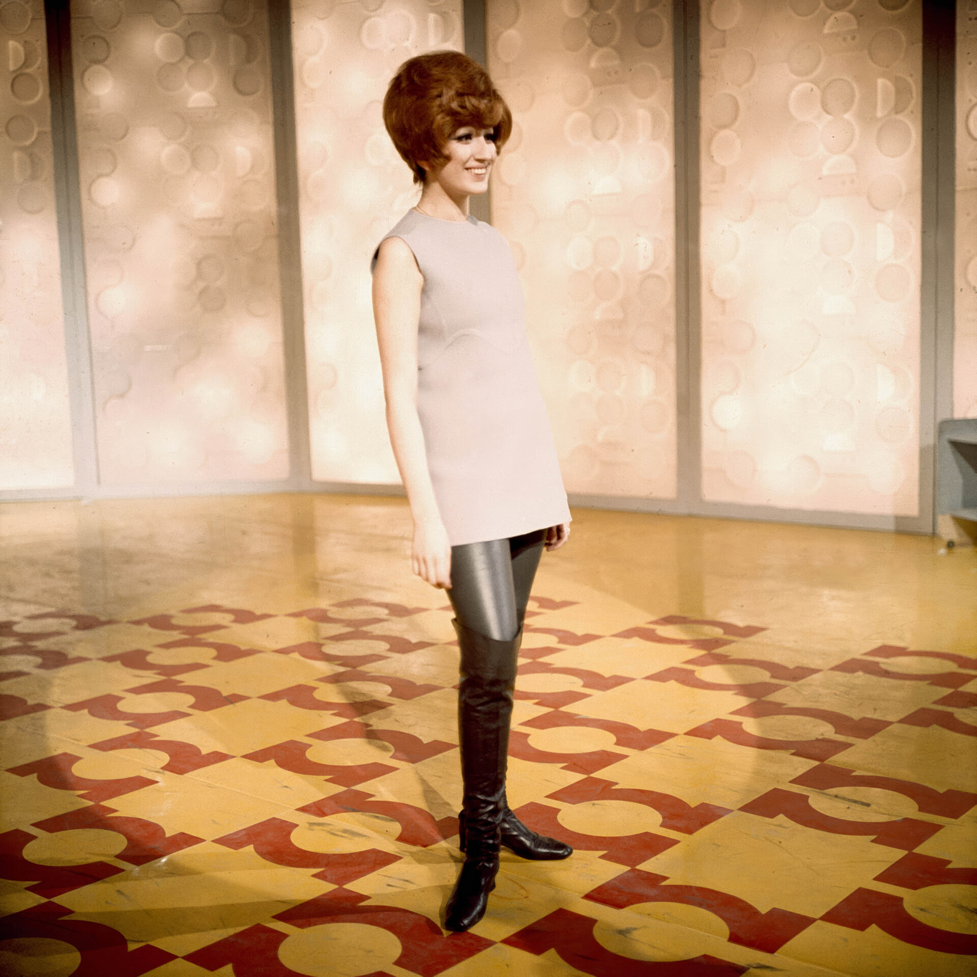 Mina's debut on RAI, she stands on a geometric-patterned floor, wearing a sleeveless top, dark pants, and knee-high boots. The background is a wall with circular designs. She smiles slightly and has a styled updo hairstyle.