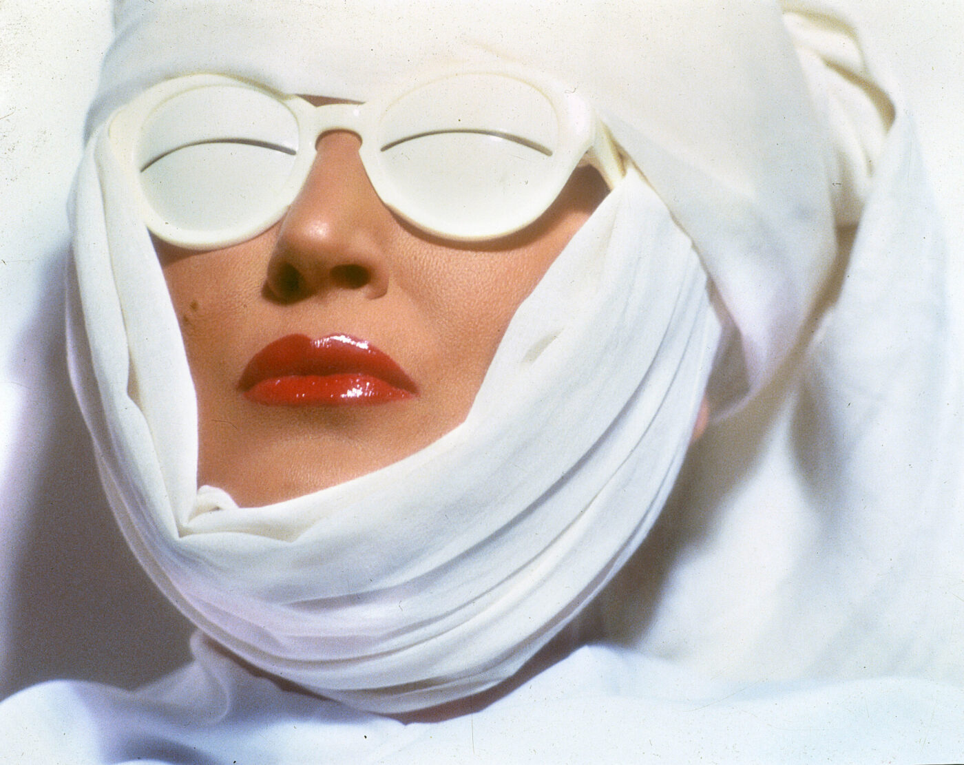 Mina with wrapped white cloth covering most of their face, wearing white sunglasses and glossy red lipstick, against a white background. The image conveys a futuristic or avant-garde aesthetic.