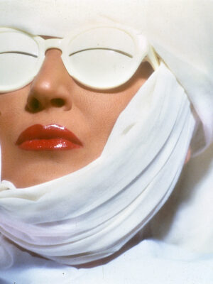 Mina with wrapped white cloth covering most of their face, wearing white sunglasses and glossy red lipstick, against a white background. The image conveys a futuristic or avant-garde aesthetic.