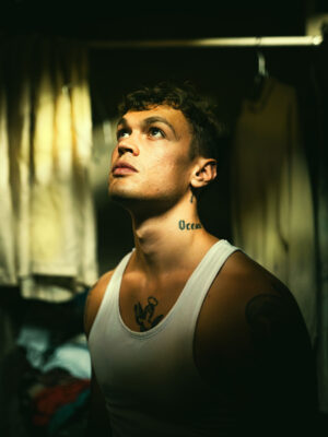 Blanco - A person with a Ocean tattoo on their neck and an anchor tattoo on their chest gazes upward. They are wearing a white tank top in a dimly lit room with hanging clothes in the background, creating a moody atmosphere.