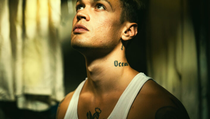 Blanco - A person with a Ocean tattoo on their neck and an anchor tattoo on their chest gazes upward. They are wearing a white tank top in a dimly lit room with hanging clothes in the background, creating a moody atmosphere.