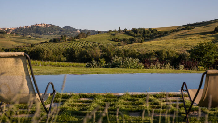 Follonico - Two lounge chairs facing a serene infinity pool overlook rolling green hills with a distant hilltop town under a clear blue sky. The lush landscape is bathed in warm sunlight, creating a peaceful and inviting atmosphere.