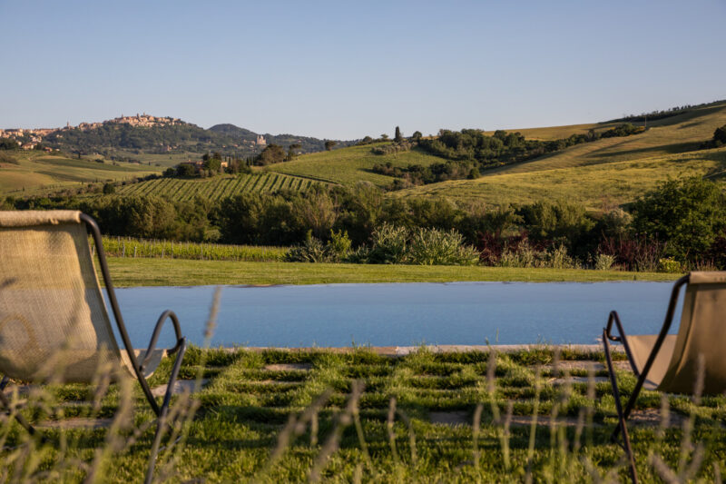 Follonico - Two lounge chairs facing a serene infinity pool overlook rolling green hills with a distant hilltop town under a clear blue sky. The lush landscape is bathed in warm sunlight, creating a peaceful and inviting atmosphere.