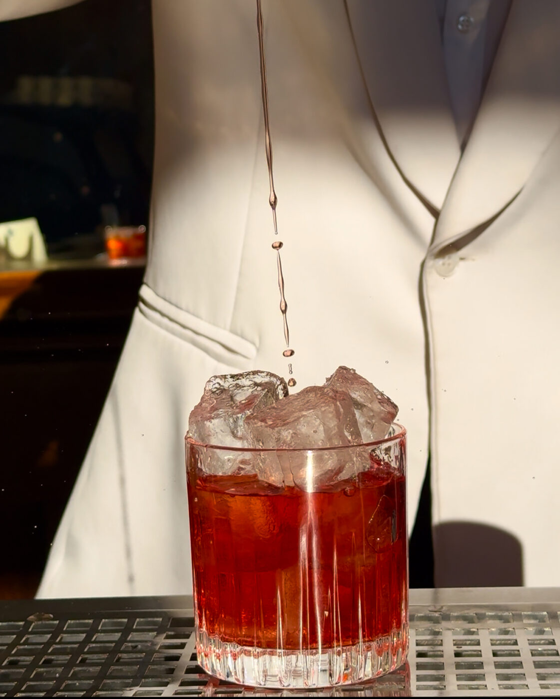 A bartender in a white jacket pours a cocktail over large ice cubes in a clear glass. The drink has a rich, dark red color.