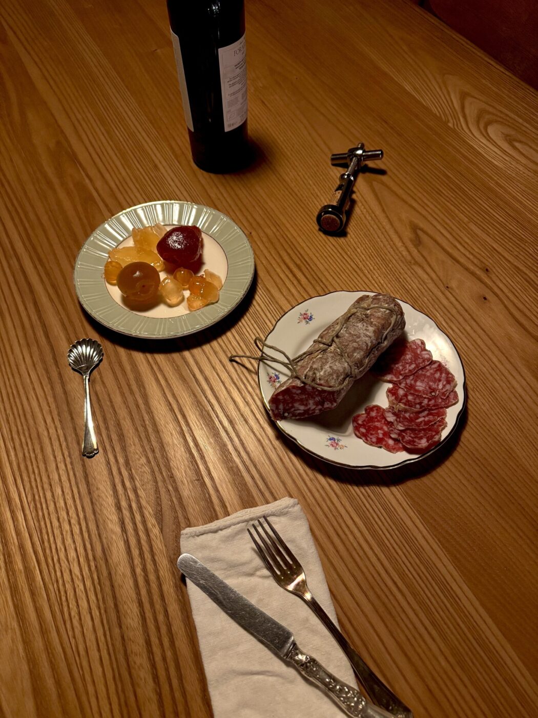 Mostarda and salame di Cremona - A wooden table setup includes a plate with sliced salami, a plate of assorted dried fruits, a bottle of wine with a corkscrew, a silver spoon, and a knife and fork placed on a napkin.