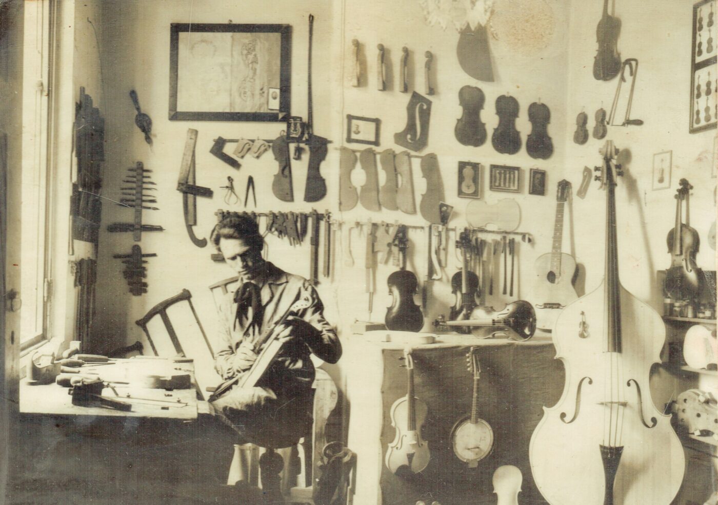 Craftsmans Haven - A sepia-toned image of a man sitting in a workshop, working on a stringed instrument. The walls are lined with various violins, guitars, and tools. Sunlight streams in through a window on the left, illuminating the workspace.