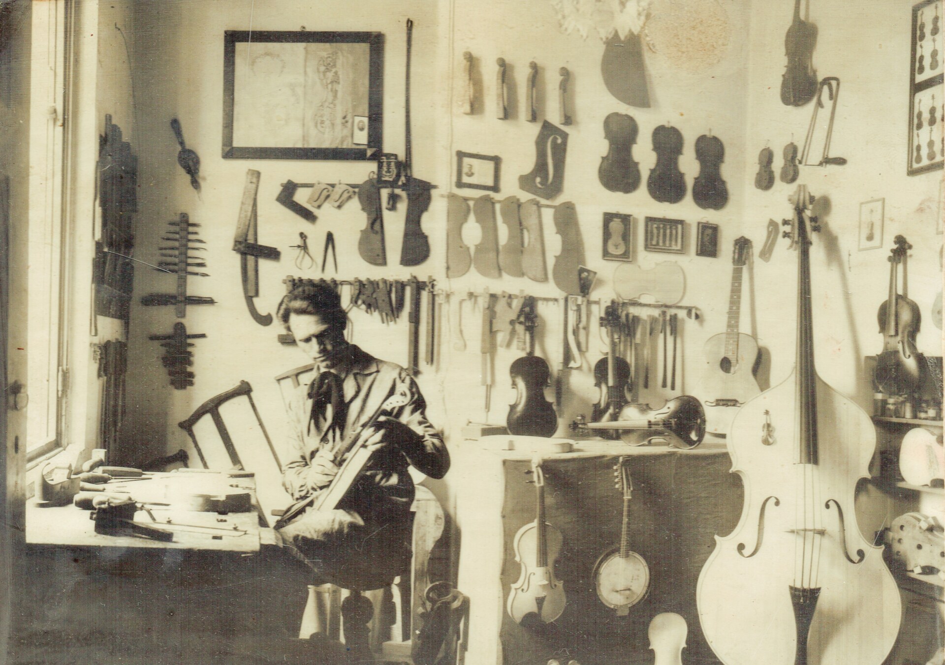 Craftsmans Haven - A sepia-toned image of a man sitting in a workshop, working on a stringed instrument. The walls are lined with various violins, guitars, and tools. Sunlight streams in through a window on the left, illuminating the workspace.