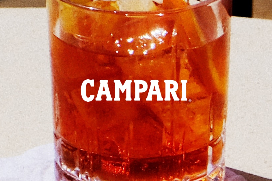 A glass of orange-red drink on a table, filled with ice cubes. The word CAMPARI is visible on the glass, indicating the brand of the beverage.