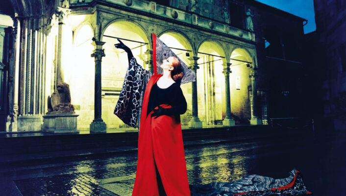 Nessuno passerà - Una persona con una lunga tunica rossa posa in modo teatrale in una strada acciottolata di notte, di fronte a un edificio storico con archi e statue. Le sue braccia sono tese con un tocco teatrale e il terreno è riflettente per la pioggia.