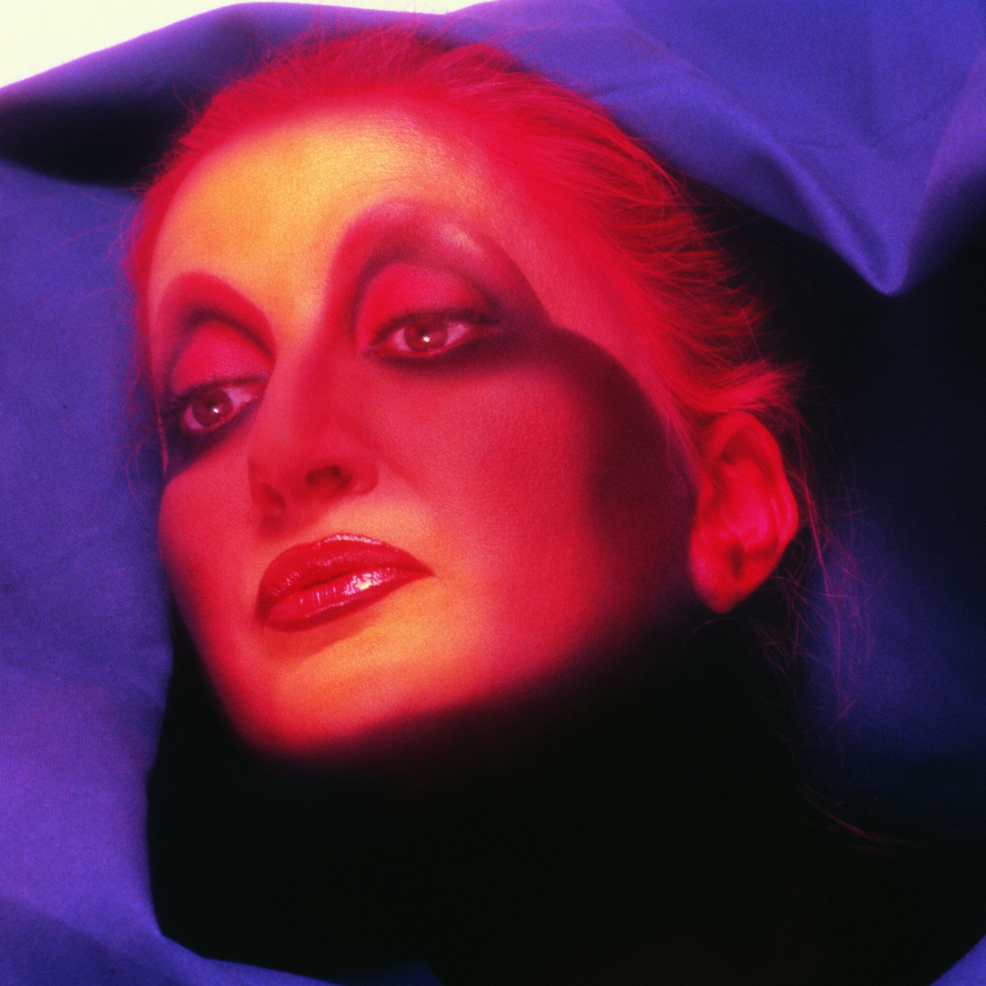 The cover of Italiana Mina. Mina with dramatic, artistic makeup and a red hue on their face is surrounded by a blue fabric backdrop. The lighting creates a high-contrast, surreal effect, highlighting glossy lips and expressive eyes.