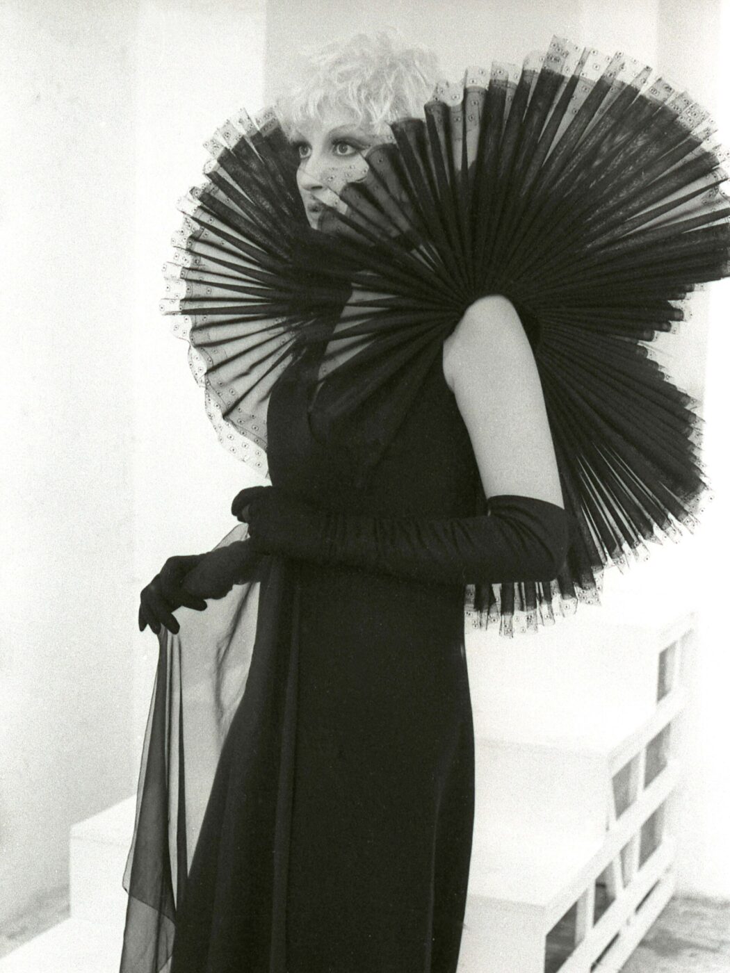 Mina with short, light-colored hair wears an elaborate black dress featuring a large, pleated collar that fans around their shoulders. They hold sheer fabric in one hand, standing against a minimalist background. For Barilla.
