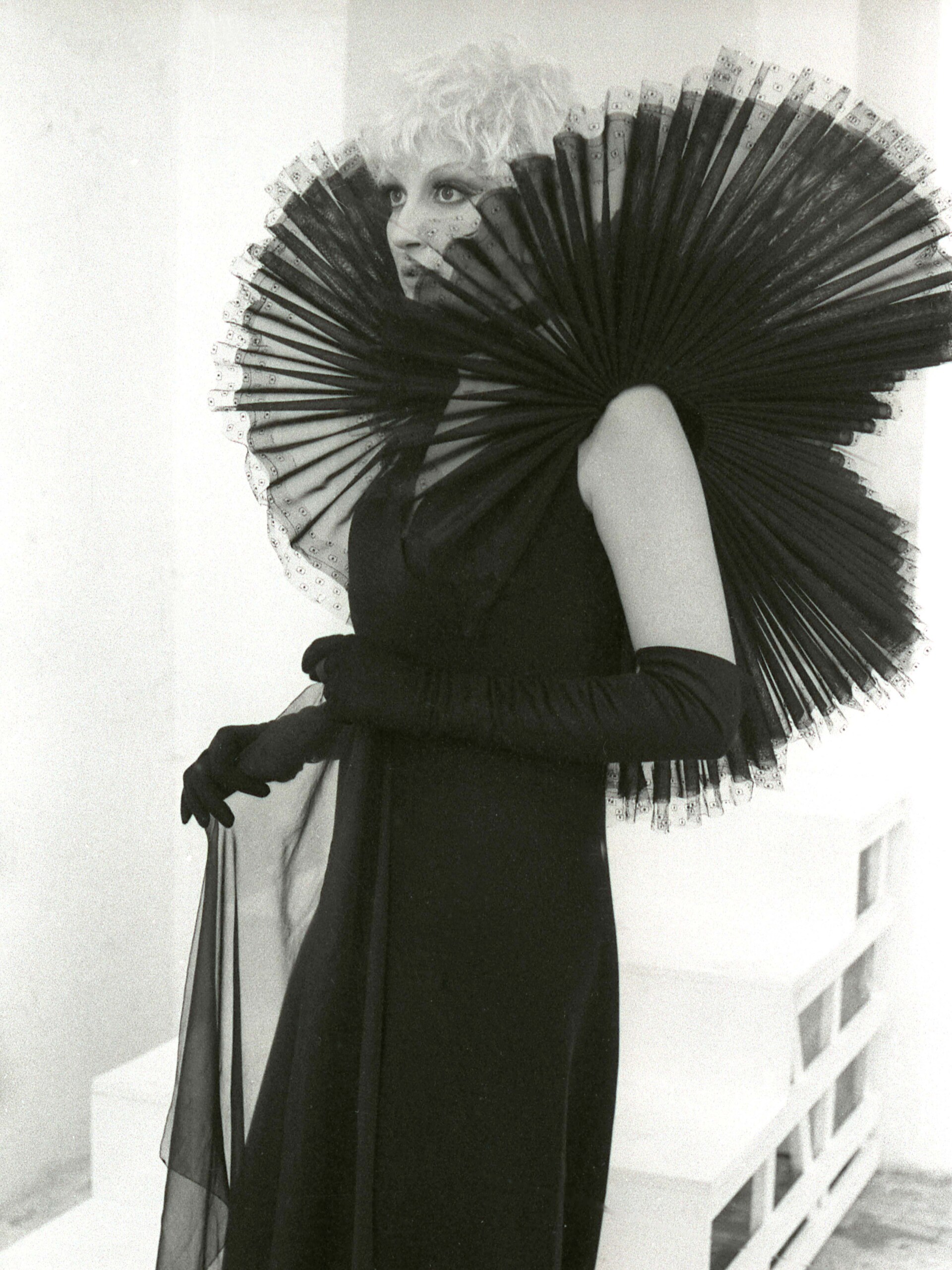 Mina with short, light-colored hair wears an elaborate black dress featuring a large, pleated collar that fans around their shoulders. They hold sheer fabric in one hand, standing against a minimalist background. For Barilla.