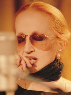 Mina wearing sunglasses and a black scarf, is seen in front of a microphone. They appear to be deep in thought or focused, with their hand near their mouth. The background is softly blurred.