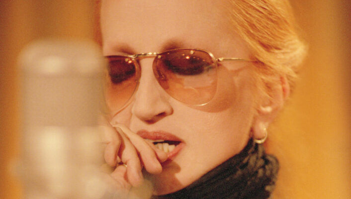 Mina wearing sunglasses and a black scarf, is seen in front of a microphone. They appear to be deep in thought or focused, with their hand near their mouth. The background is softly blurred.