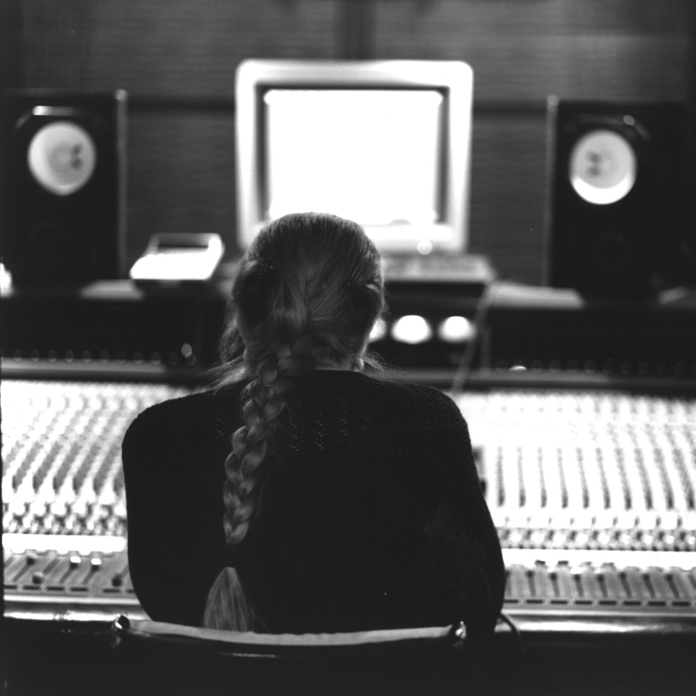 Mina with a long braid sits facing a recording studio mixing console. There are large speakers on either side of a computer monitor displaying a bright screen in the dimly lit room.