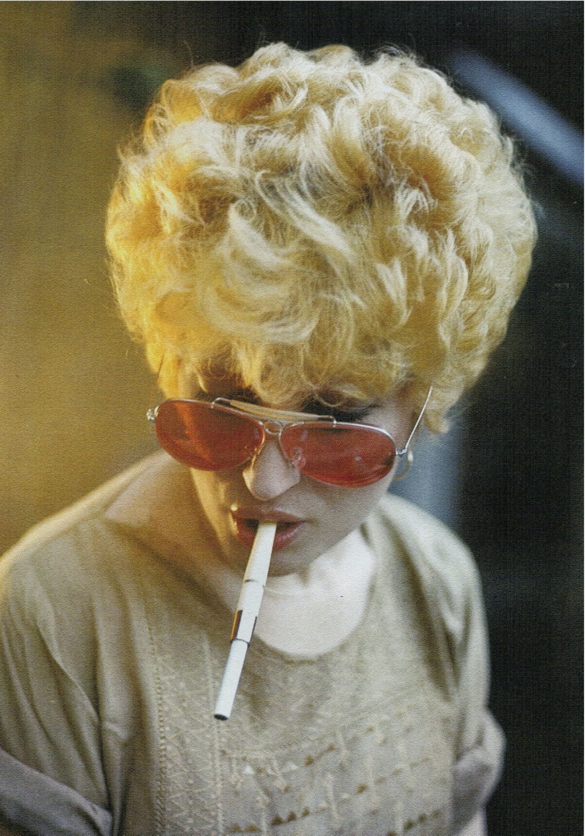 Mina Mazzini with voluminous, curly blond hair wears red-tinted sunglasses and holds a cigarette in their mouth. They are wearing a light-colored top and are looking downward. The image has a vintage feel.