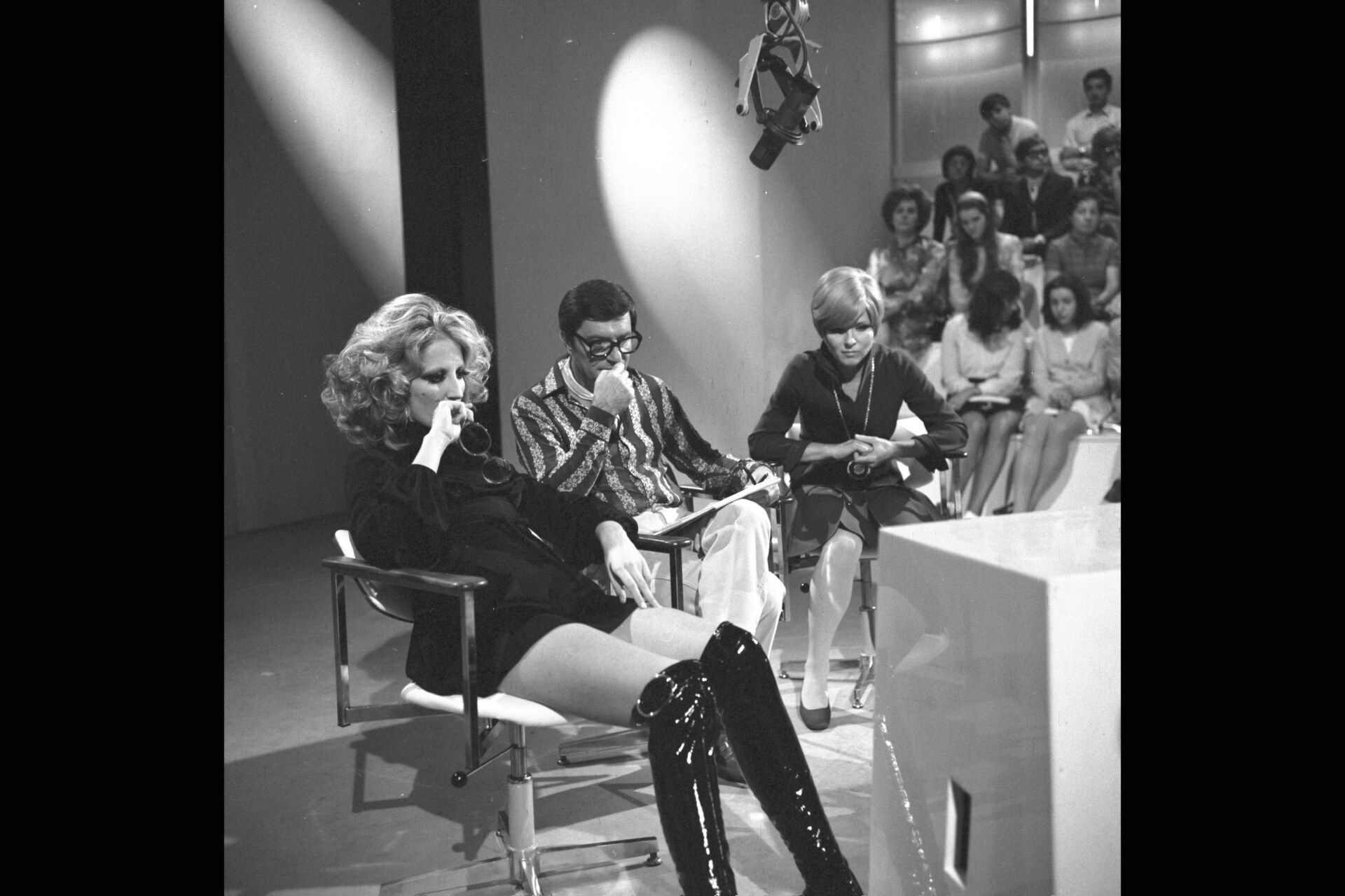 Mina backstage at RAI - Black and white photo of three people seated on a stage during a television show. The person on the left wears a dress and thigh-high boots, the person in the middle wears glasses and a striped shirt, and the person on the right is wearing a light jacket. An audience is visible in the background.