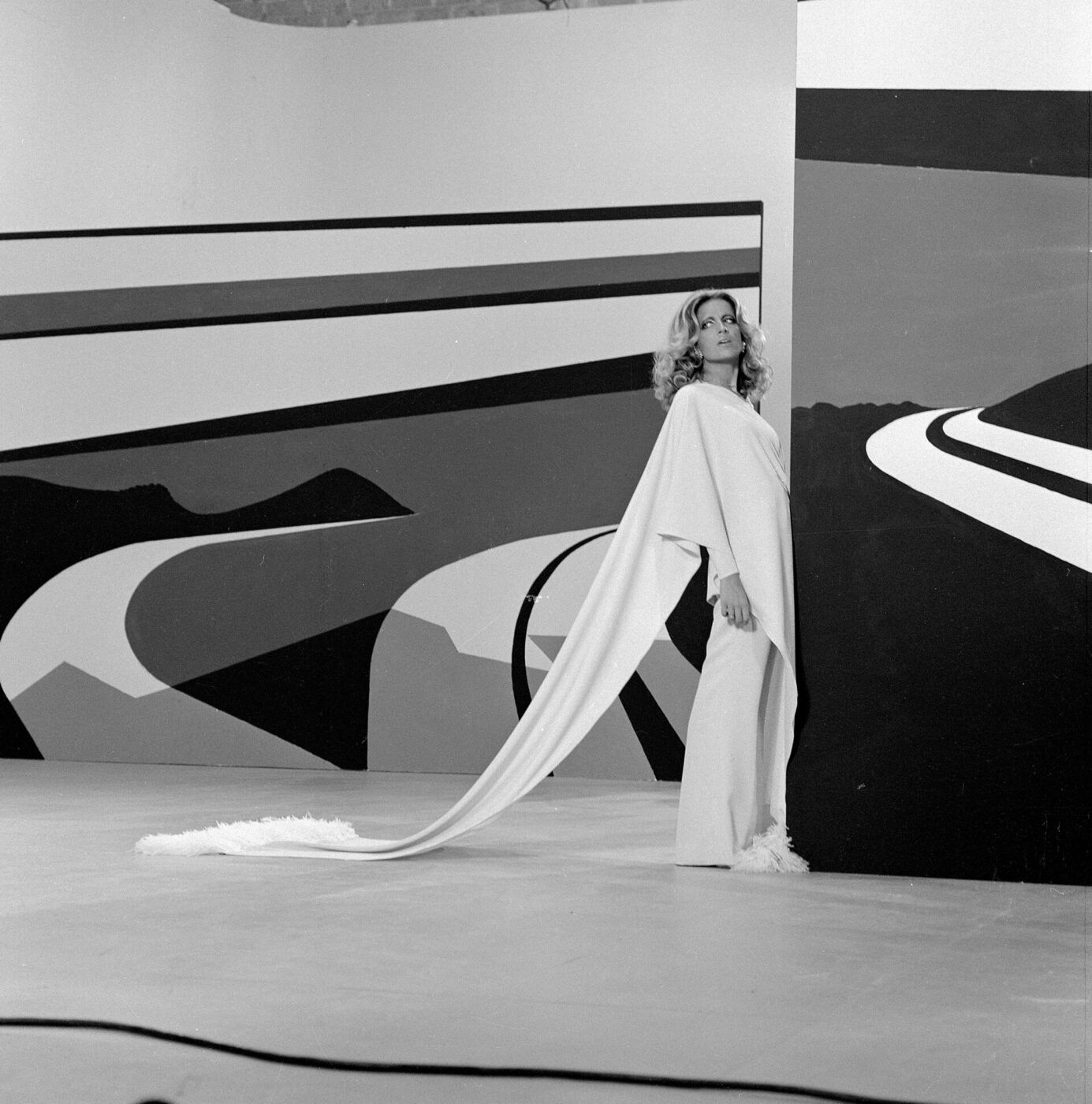 Mina stands beside an abstract mural wearing a long, flowing garment. The artwork features bold geometric patterns in black, white, and gray. The scene is set in a minimalistic indoor space, and she gazes thoughtfully ahead for a Barilla commercial