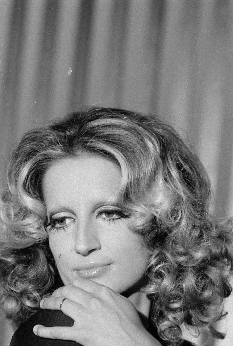 Black and white portrait of Mina with wavy, shoulder-length hair. They rest their chin on their hand, looking thoughtfully to one side. The background is softly blurred with vertical lines.