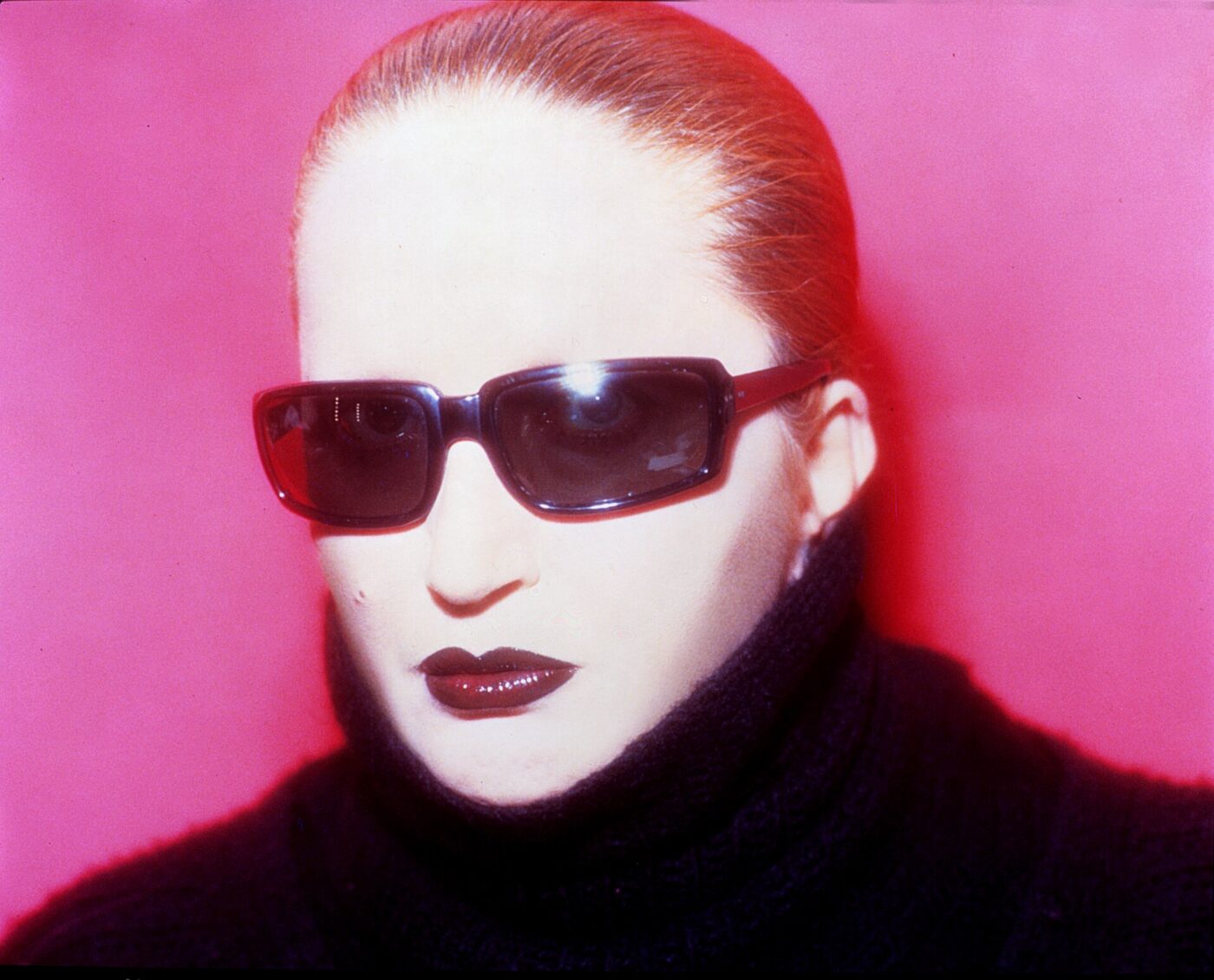 Mina Mazzini wearing dark sunglasses and a black turtleneck against a bright pink background. Their hair is slicked back, and they have bold makeup with dark lipstick.