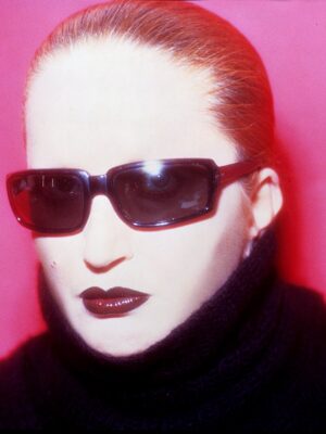 Contrasto elegante - Una persona con i capelli lisci e lisci, occhiali da sole scuri e rossetto audace, è su uno sfondo rosa acceso. Indossa un maglione a collo alto nero, creando un look drammatico ed elegante.