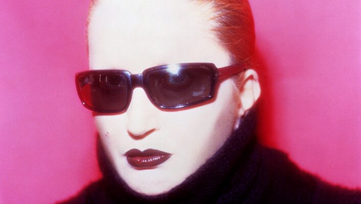 Mina Mazzini wearing dark sunglasses and a black turtleneck against a bright pink background. Their hair is slicked back, and they have bold makeup with dark lipstick.