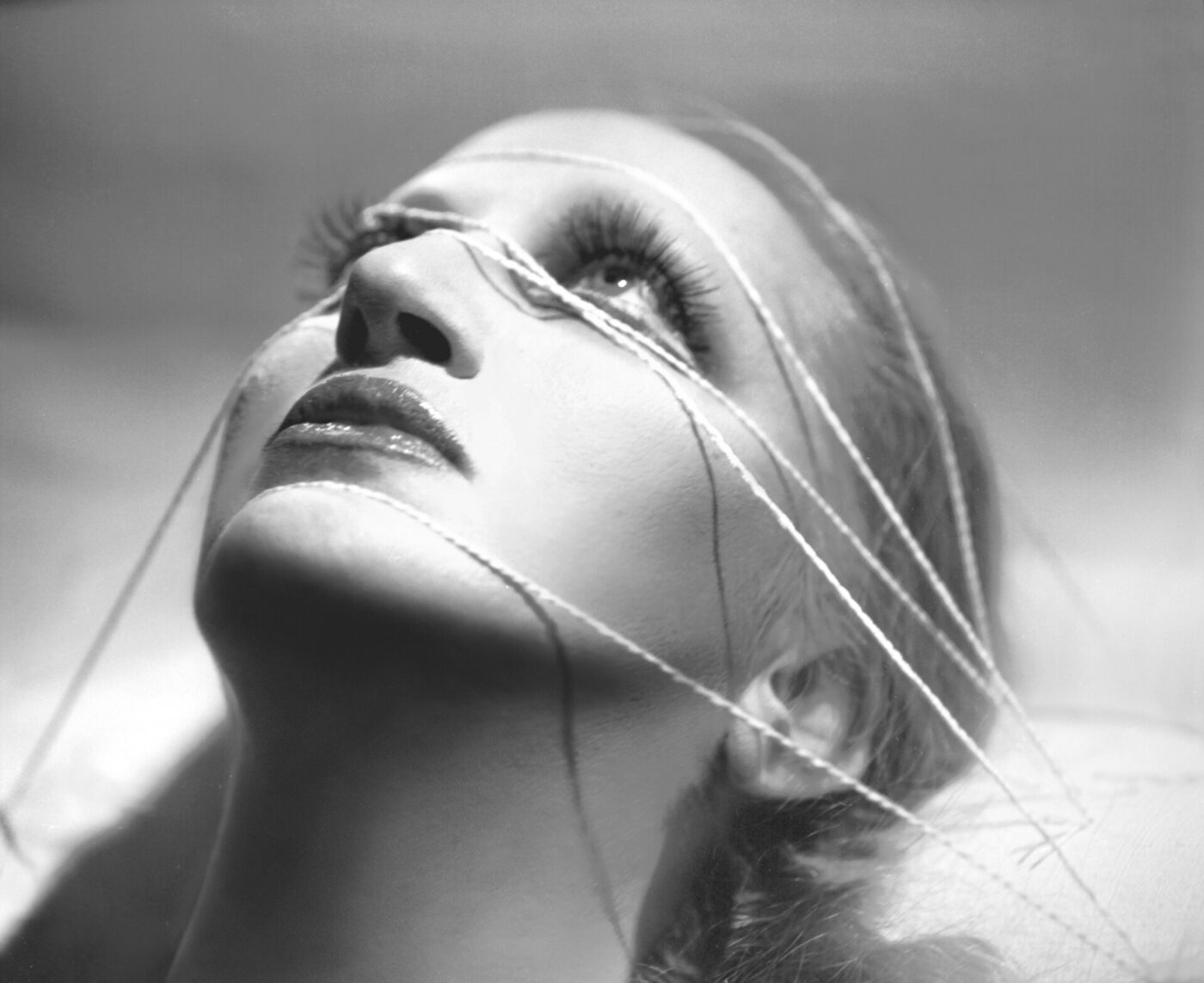 Mina's album cover encadenados - Black and white photo of singer Mina looking upward, face partially covered with strands of string. The strings create geometric patterns across the face, highlighting the elongated eyelashes and glossy lips. The lighting emphasizes the contours of the face.