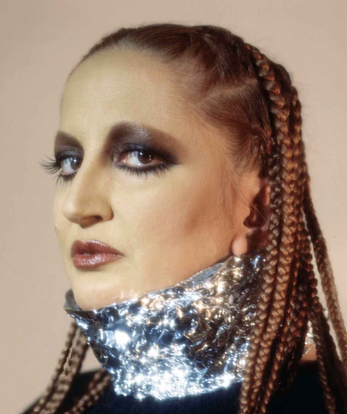 Futuristic Glam - Singer Mina with braided hair and dramatic makeup wears a metallic, reflective collar. The makeup features bold, smoky eyeshadow and glossy lips. The background is a soft beige, and the person gazes slightly to the side.